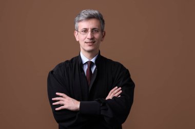 Kahverengi arka planda çapraz kolları olan mahkeme kıyafetli gülümseyen yargıç.