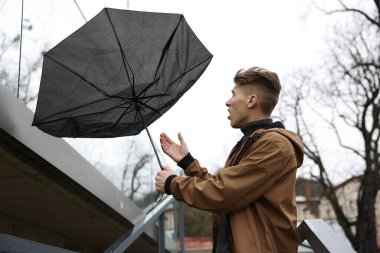 Kırık şemsiyeli bir adam açık havada yağmur altında rüzgarla savaşıyor.