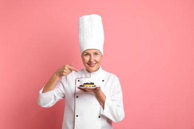 Profesyonel pasta şefi pembe arka plandaki tatlıyı işaret ediyor.