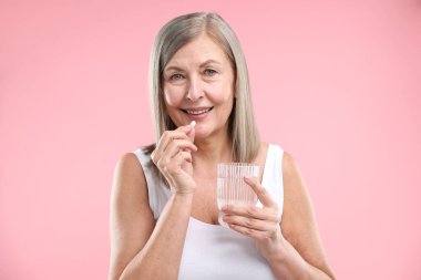 Bir bardak suyla gülümseyen kadın pembe arka planda kolajen hapı alıyor.