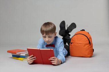 Sırt çantası açık gri arka planda kitap okuyan sevimli bir okul çocuğu.