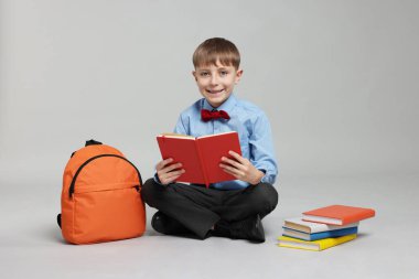 Sırt çantası açık gri arka planda kitap okuyan sevimli bir okul çocuğu.