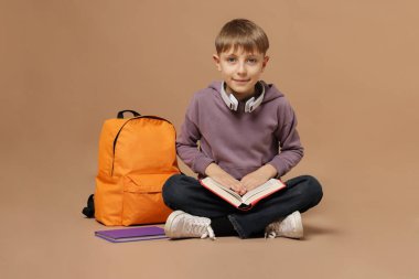 Kulaklıkları ve sırt çantasıyla kahverengi arka planda kitap okuyan tatlı bir okul çocuğu.