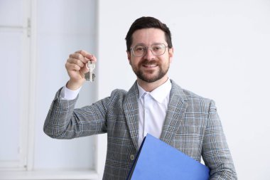 Emlakçının elinde ev şeklindeki anahtarlık ve klasör var.