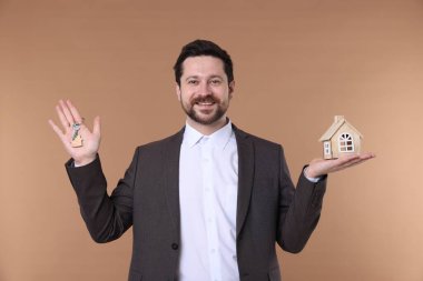 Emlakçının elinde anahtarlık ve bej arka planda ev modeli var.