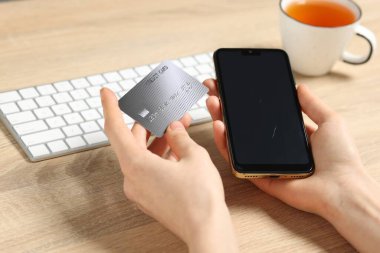 Kredi kartı ve akıllı telefonu olan bir kadın ahşap masada internetten ödeme yapıyor.