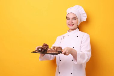 Üniformalı şekerlemeci turuncu arka planda lezzetli çikolatalı tatlılar tutuyor, mesaj için yer var.