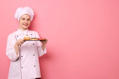 Üniformalı şekerlemeci pembe arka planda farklı lezzetli tatlılar tutuyor, mesaj için yer var.