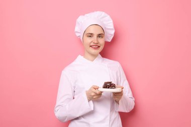 Üniformalı şekerci pembe arka planda çikolatalı tatlı tutuyor.