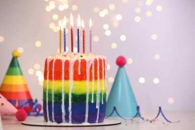 Enfes gökkuşağı doğum günü pastası, yanan mumlar, flamalar, serpiştirmeler ve parti şapkaları beyaz masanın üzerinde bulanık ışıklı arka planda, yakın plan. Metin için boşluk