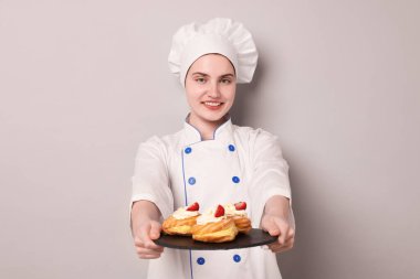 Üniformalı şekerci açık gri arka planda lezzetli profiteroller tutuyor.