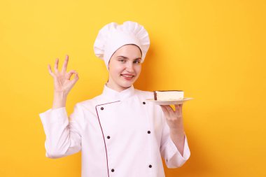 Üniformalı bir şekerci elinde lezzetli bir pasta tutuyor ve turuncu arka planda güzel bir jest yapıyor.