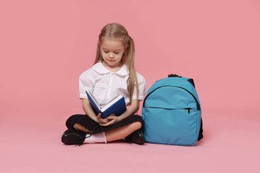 Sırt çantalı liseli kız pembe arka planda kitap okuyor.
