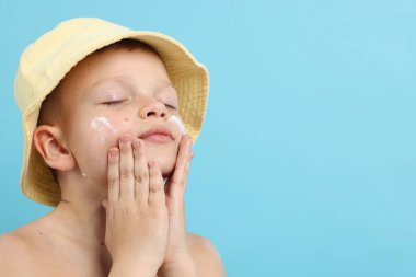 Küçük çocuk açık mavi arka planda cildine güneş kremi sürüyor.