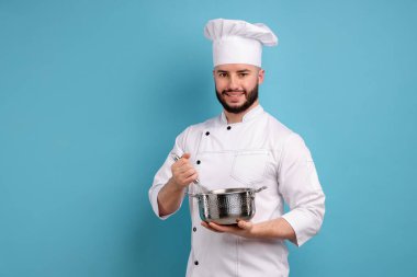 Üniformalı mutlu pastacı elinde çırpıcı ve açık mavi arka planda pişirme kabı tutuyor.