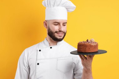 Üniformalı mutlu pastacı turuncu arka planda çikolatalı pasta tutuyor.