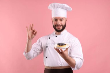 Üniformalı mutlu pastacı elinde yaban mersinli turta ve pembe arka planda güzel bir jest gösteriyor.