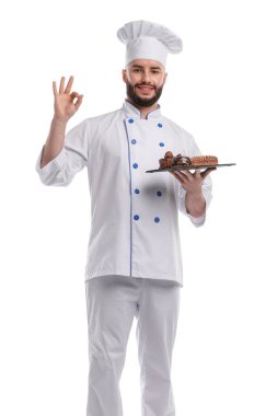 Üniformalı mutlu pastacı lezzetli çikolatalı tatlılar tutuyor ve beyaz arka planda iyi bir jest yapıyor.