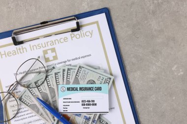 Sağlık sigortası kartı, sigorta poliçesi, para, gözlükler ve kalemler gri masada.