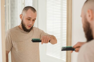 Kellik sorunu. Saç çizgisi gerilemiş bir adam aynanın yanında fırça tutuyor.