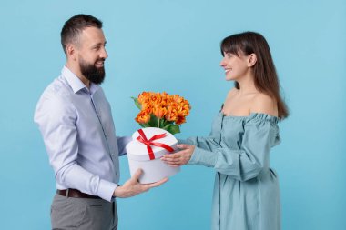 Bir erkek, açık mavi arka planda sevgili karısına güzel laleler ve hediye kutusu sunar.