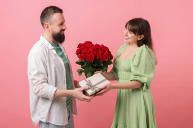 Kırmızı gül buketini ve hediye kutusunu pembe arka plandaki sevgilisine sunan bir adam.