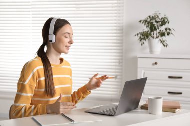 İçerideki masada bilgisayardan öğretmenle çevrimiçi ders alan gülümseyen kadın.