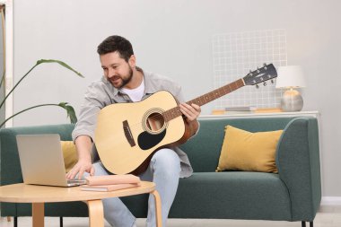 Rahatlatıcı hobi. Evdeki kanepede dizüstü bilgisayarla gitar çalan gülümseyen adam.