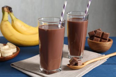 Bardaklarda çikolatalı süt, parçalar ve muzlar mavi ahşap masada, yakın plan.