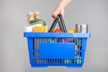 Elinde gri arka planda farklı ürünler olan alışveriş sepeti tutan adam, yakın plan.