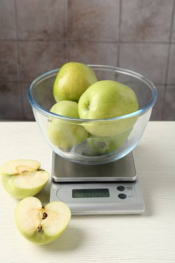 Electronic kitchen scale with bowl of green apples on white wooden table against tiled wall