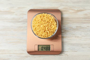 Modern elektronik mutfak terazisi ve ahşap masa üzerinde bir kase çiğ makarna.