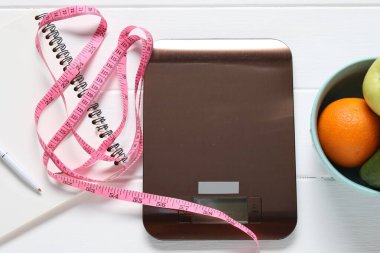 Elektronik mutfak ölçeği, mezura, not defteri ve beyaz ahşap masa üzerindeki ürünler, düz zemin. Kalori kontrolü