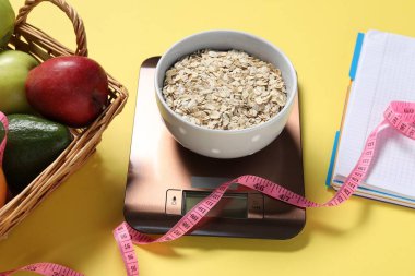 Elektronik mutfak ölçeği, mezura, not defteri ve sarı arka plandaki ürünler, yakın plan. Kalori kontrolü