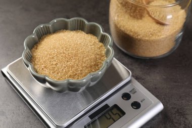 Dijital mutfak terazisi, gri masanın üzerinde kahverengi şeker kasesi, yakın plan.