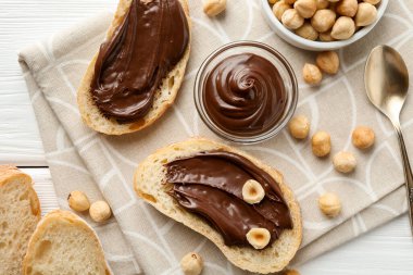 Çikolata sürülmüş ekmek dilimleri ve beyaz ahşap masada fındıklar, düz uzanmış.