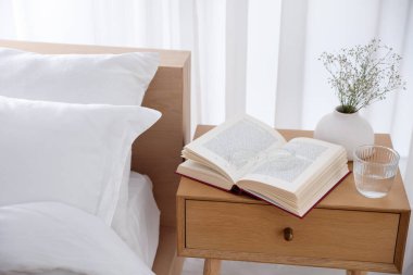 İçinde kitap olan komodin, vazoda çiçekler, yatağın yanında bir bardak su ve bardaklar.