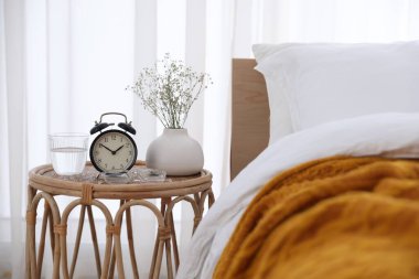 Komodinin yanında çalar saat, vazoda çiçekler, yatağın yanında bir bardak su ve bardaklar.