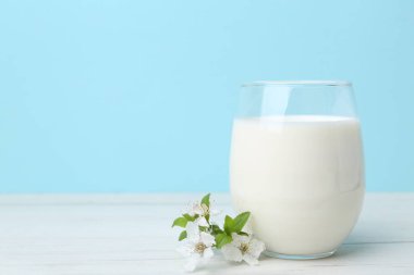 Açık mavi arka planda beyaz ahşap masada bir bardak taze süt ve çiçekler. Metin için boşluk