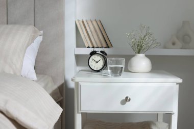Komodinin yanında çalar saat, vazoda çiçekler ve yatağın yanında bir bardak su.
