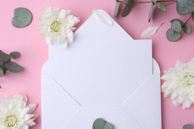 Kağıt zarftaki boş tebrik kartı, kasımpatı çiçekleri ve pembe arkaplanda okaliptüs dalları, düz uzanmış.