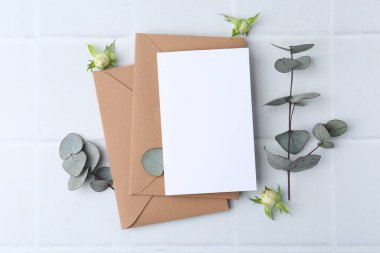 Boş tebrik kartı, kağıt zarflar, gül çiçekleri ve okaliptüs dalları ışık kareli zemin üzerinde, düz uzanmış