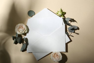 Boş tebrik kartı, zarflar, güller ve okaliptüs yaprakları bej arka planda, düz uzanmış