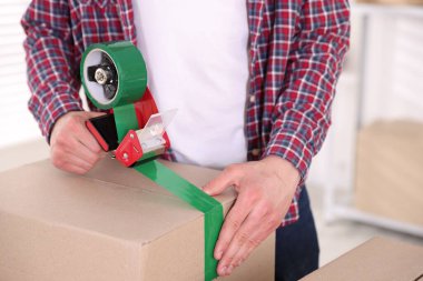 Karton kutuyu kapalı bir yere kapatmak için yapıştırıcılı bant kullanan bir adam.
