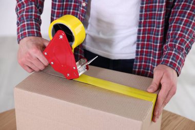 Karton kutuyu kapalı bir yere kapatmak için yapıştırıcılı bant kullanan bir adam.