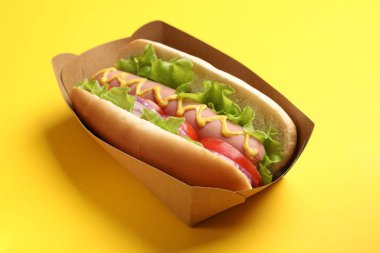 Sarı arka planda domatesli, soğanlı, marullu ve hardallı lezzetli sosisli, yakın plan.