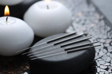 Akupunktur iğneleri, spa taşı ve koyu desenli arka planda yanan mumlar, yakın plan.