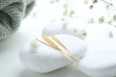 Akupunktur iğneleri, çakıl taşları ve beyaz desenli masa üzerinde çiçekli dallar, yakın plan.