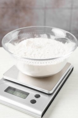 Electronic kitchen scale with bowl of flour on white wooden table against tiled wall, closeup