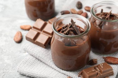 Cam kavanozlarda lezzetli puding, çikolata ve kakao çekirdekleri açık gri masada, yakın plan.